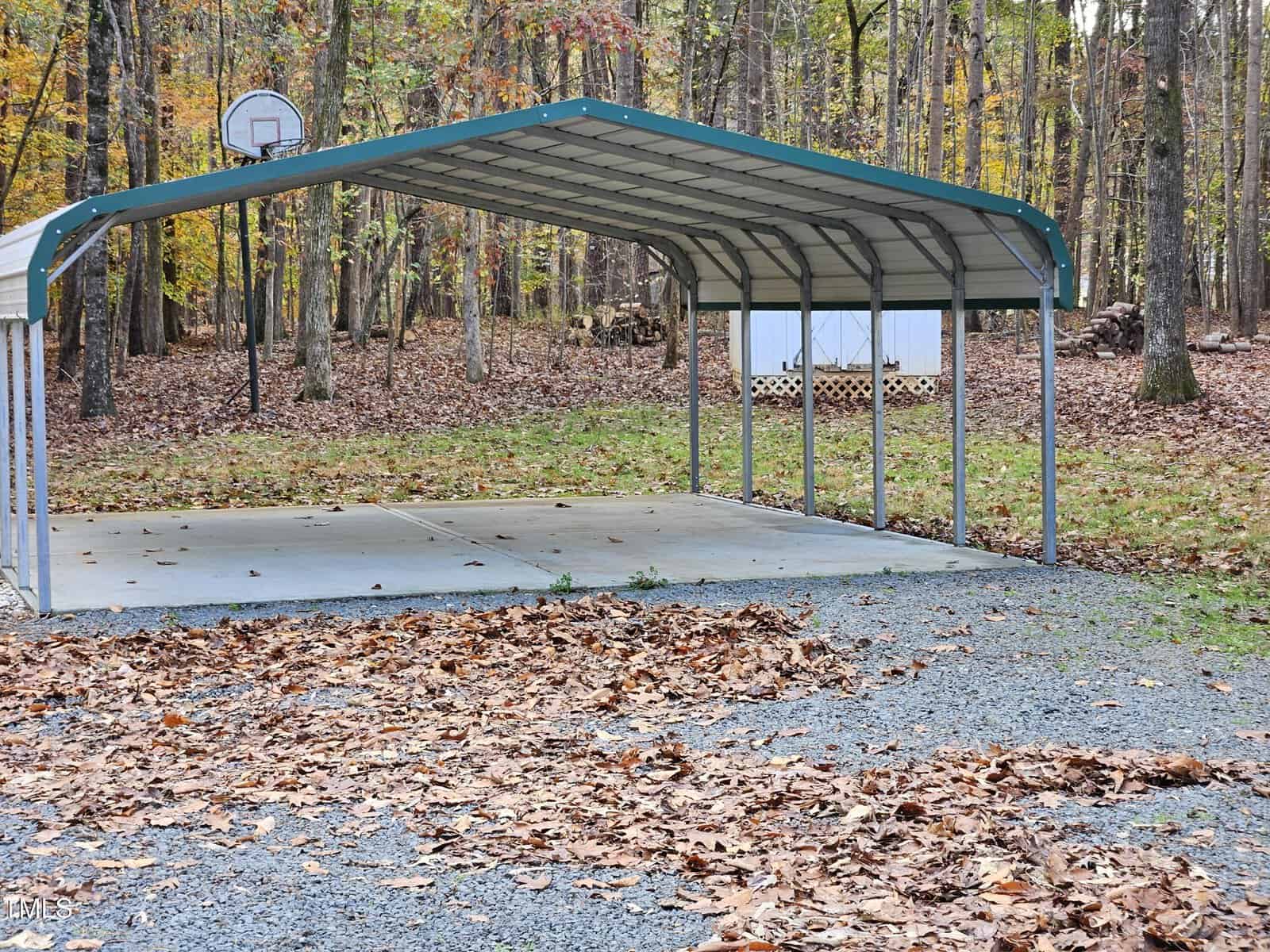 A metal carport with a green roof stands on a concrete slab in a wooded area. Fallen leaves cover the ground around it, and a basketball hoop is attached to the back of the structure. A small white shed is visible in the background.