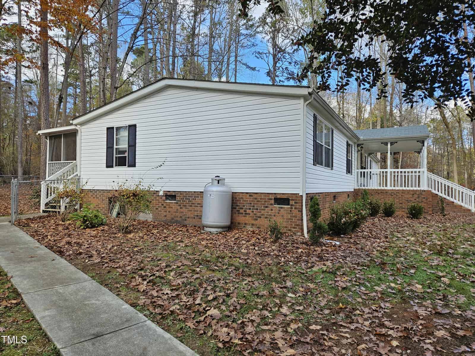A white, single-story manufactured home with brick foundation sits in a wooded area. A concrete path leads to the entrance, and fallen leaves cover the surrounding yard. A propane tank is positioned next to the home in the side yard.