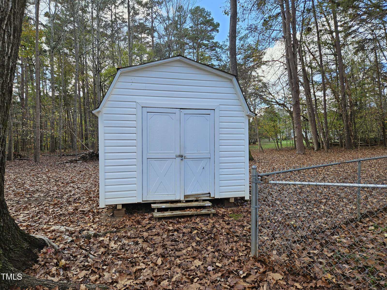 A white storage shed with double doors stands in a wooded area, surrounded by fallen leaves. A chain-link fence is partially visible in the foreground, and tall trees are in the background under a partly cloudy sky.