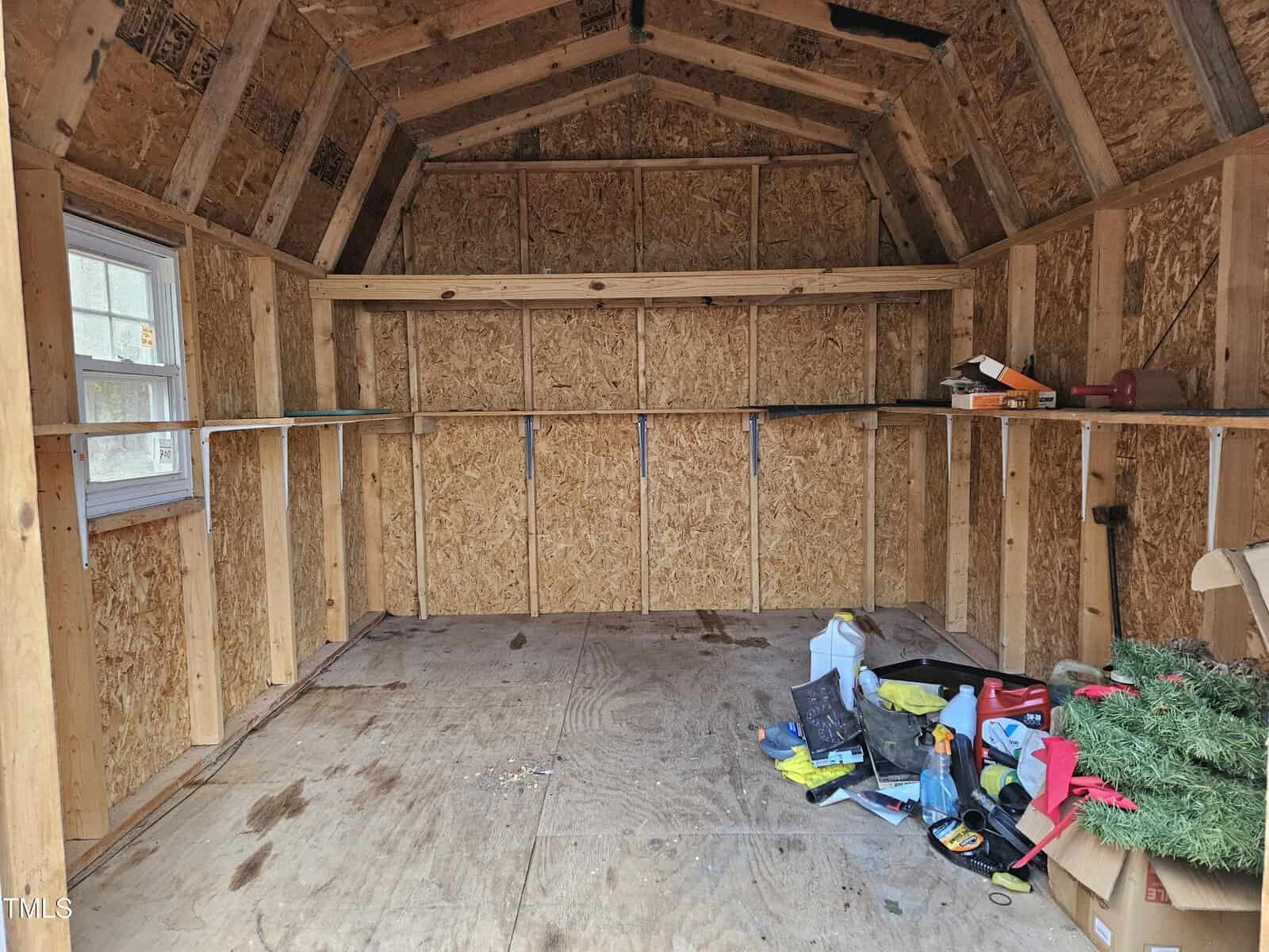 Interior of an unfinished wooden shed with plywood walls and ceiling. A window is on the left, and a shelf runs along the back wall. Various items, including tools and a piece of artificial greenery, are scattered on the floor.