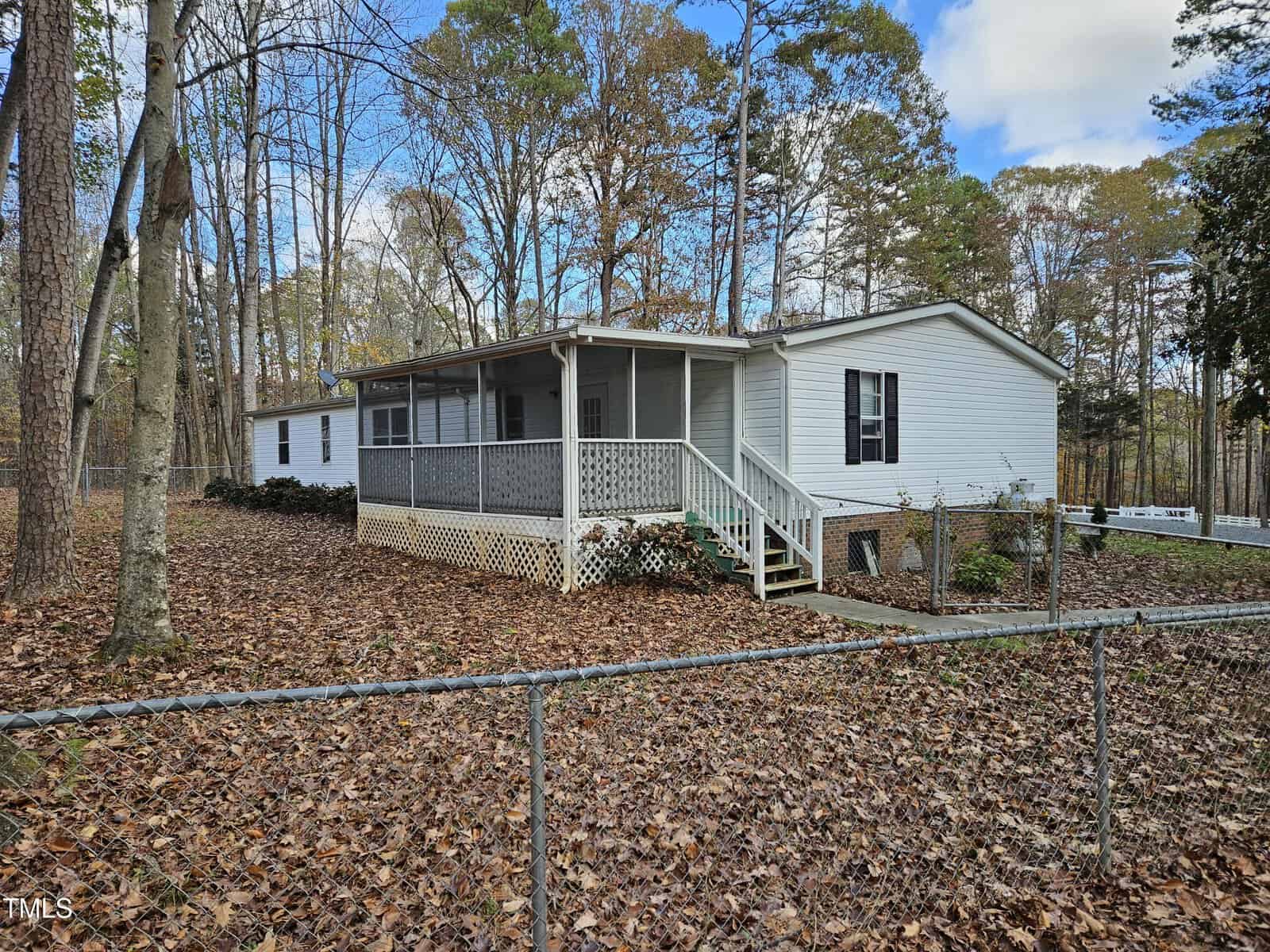 Single-story white mobile home with a covered front porch and lattice skirting, set amidst tall trees with autumn leaves covering the ground. A chain-link fence surrounds the property, and the sky is partly cloudy.