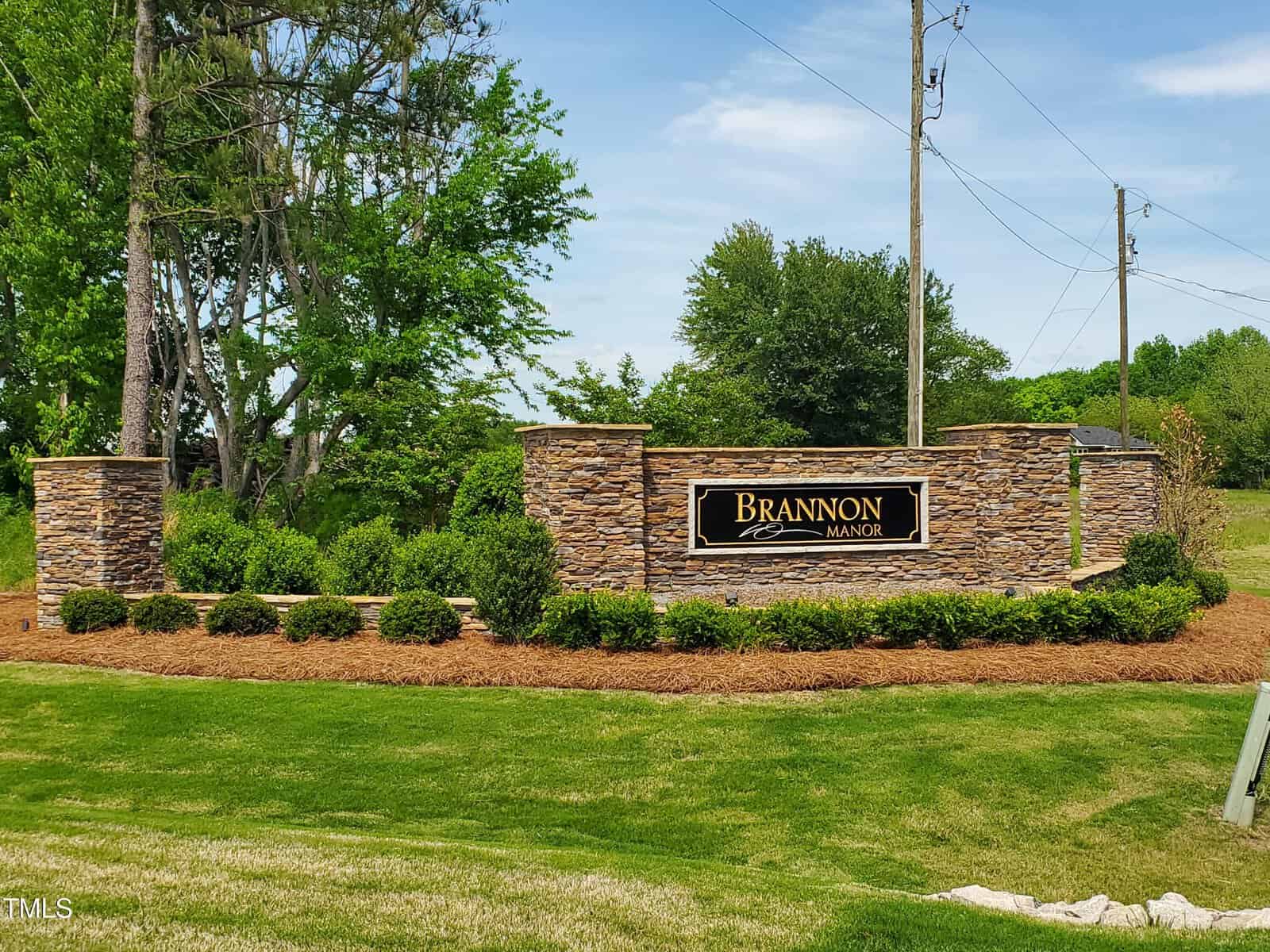 A stone entrance sign for Brannon Manor is surrounded by neatly trimmed bushes, mulch, and green grass. Trees and power lines are visible in the background under a blue sky.