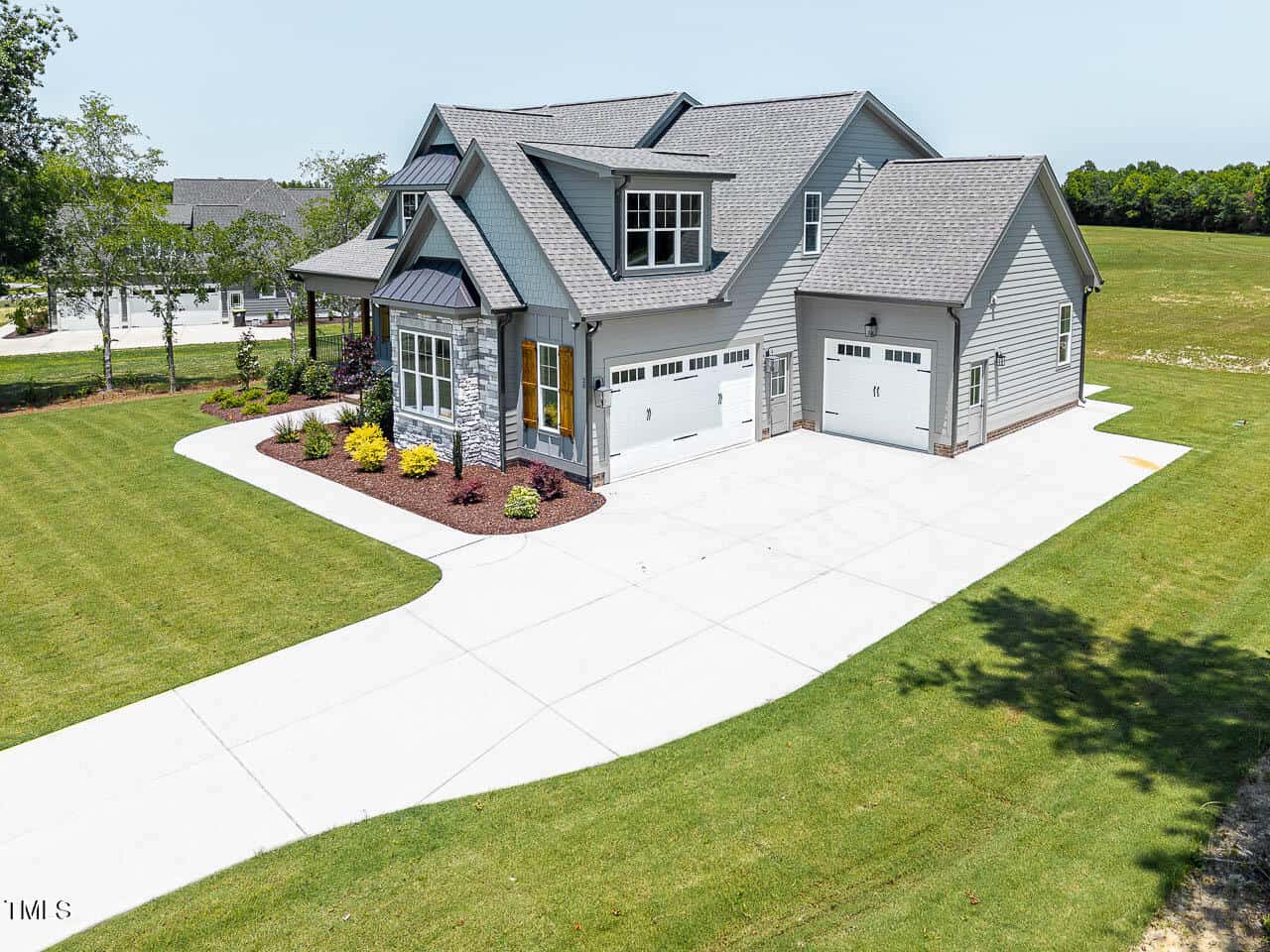 Modern two-story gray house with three-car garage, landscaped front yard, and large concrete driveway, set on a spacious green lawn with trees in the background on a sunny day.