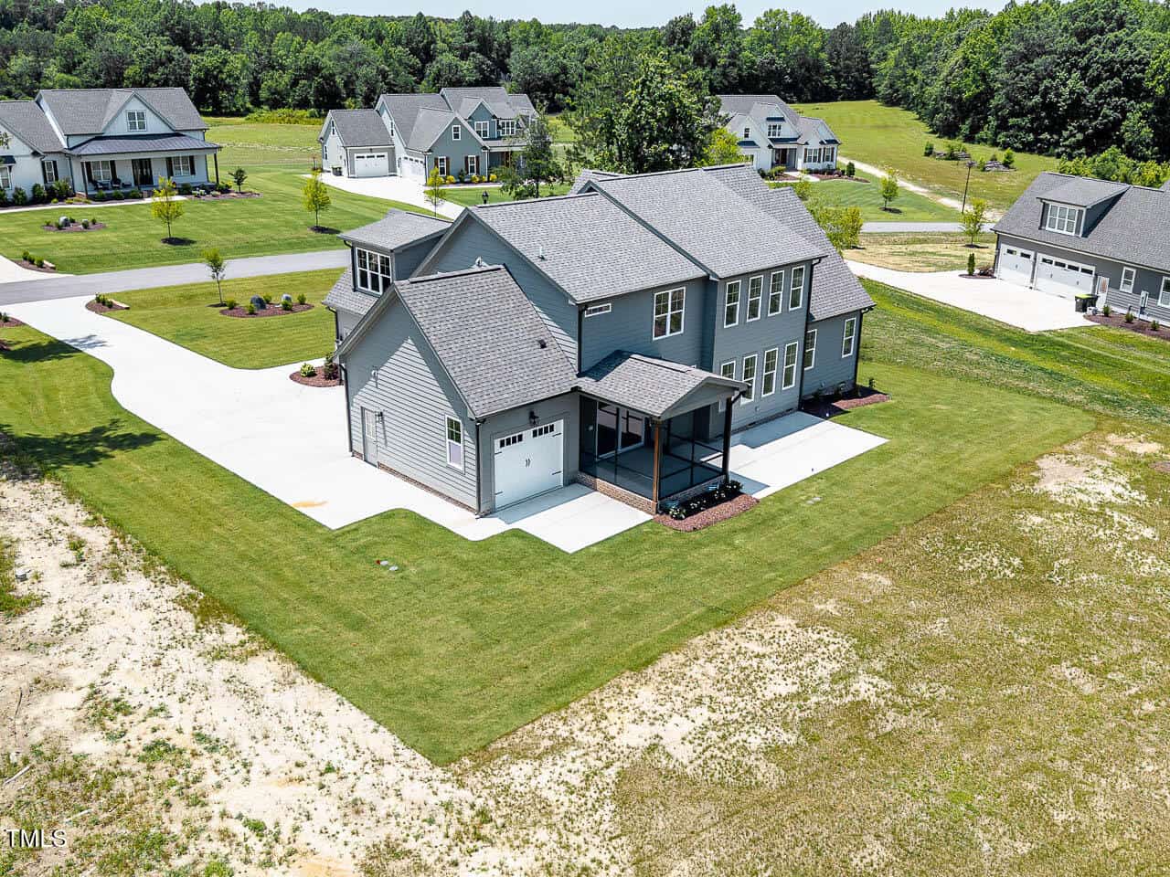 A large two-story gray house with a screened porch, attached garage, and driveway sits on a spacious grassy lot in a suburban neighborhood with several similar homes and surrounding green trees.