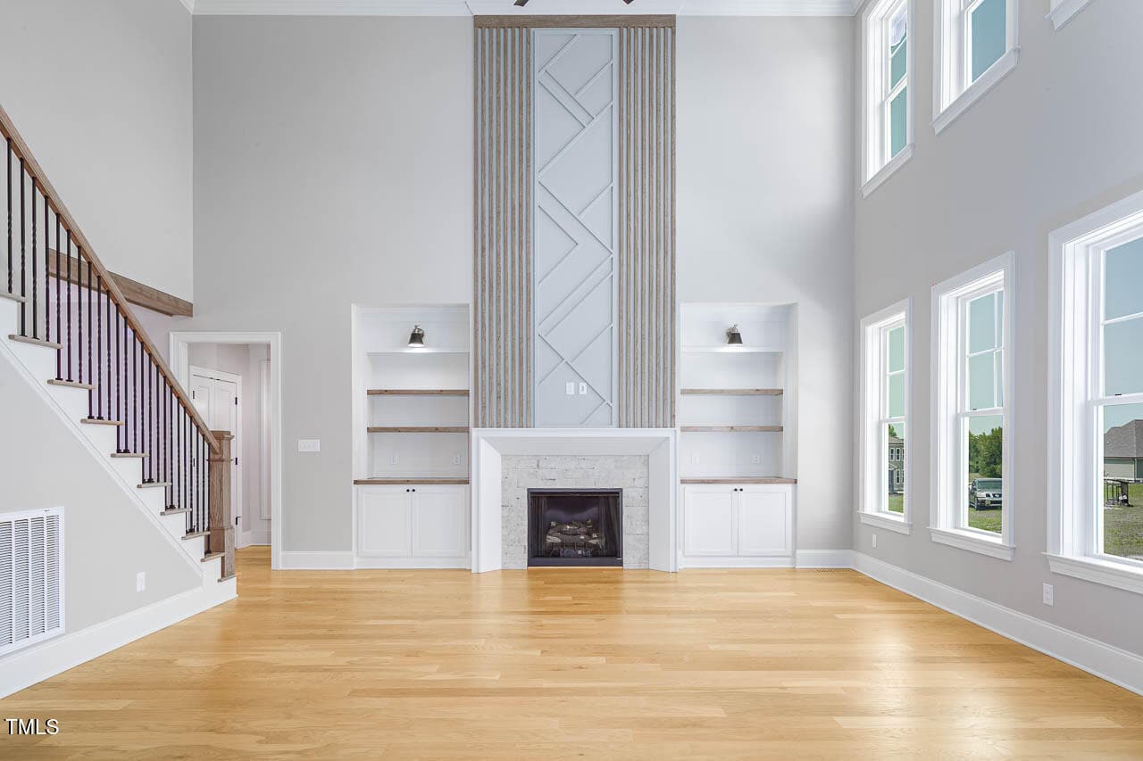 A modern, bright living room with light wood floors, a fireplace centered between built-in shelves, tall ceilings, and large windows letting in natural light. Stairs with a wooden banister are on the left.