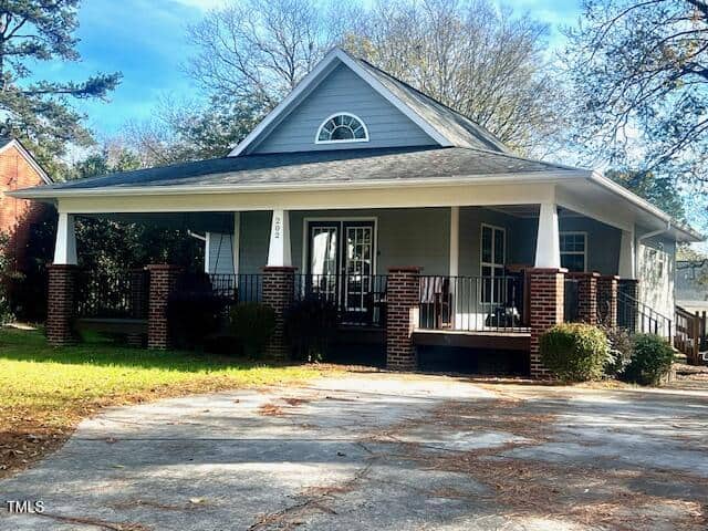 Located on 202 Delacroix Street in Oxford, this gray single-story house features a charming triangular attic window and a wraparound porch supported by white columns on brick bases. The front yard boasts a concrete driveway, lush grass, and is enveloped by trees and shrubs.