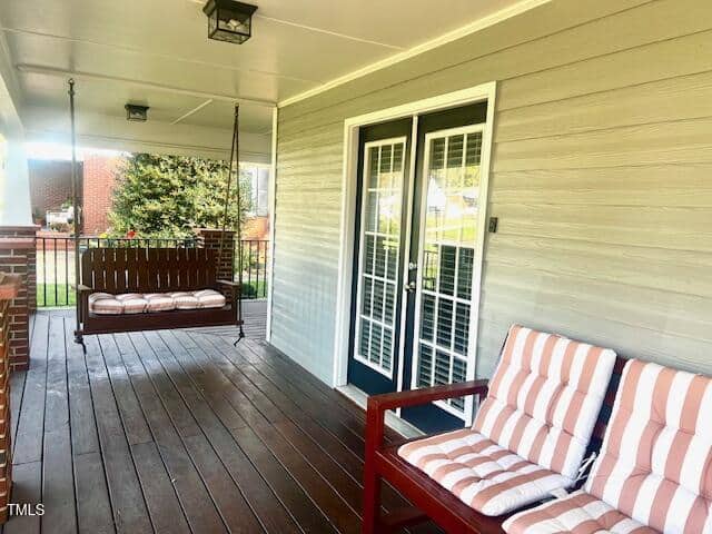 A wooden porch swing hangs next to cushioned chairs on a covered porch. The porch has a wooden floor and leads to double doors with glass panels. A brick pillar is visible on the left side, and there is greenery in the background.