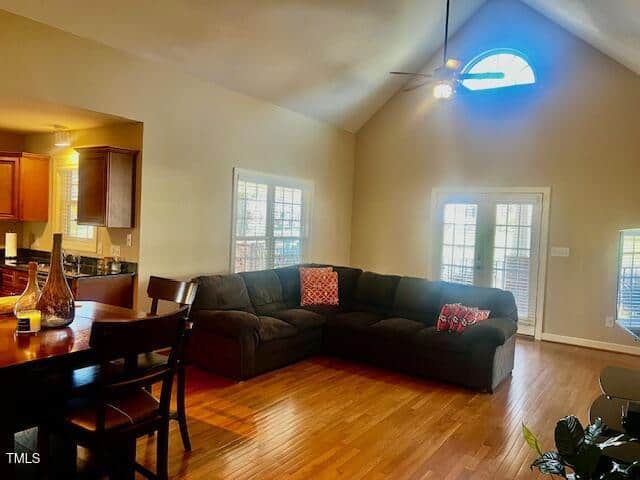 A living room with a large dark sectional sofa adorned with red and orange patterned cushions. The room features a vaulted ceiling, wooden flooring, a round wooden dining table, and natural light coming through windows and a glass-paned door.