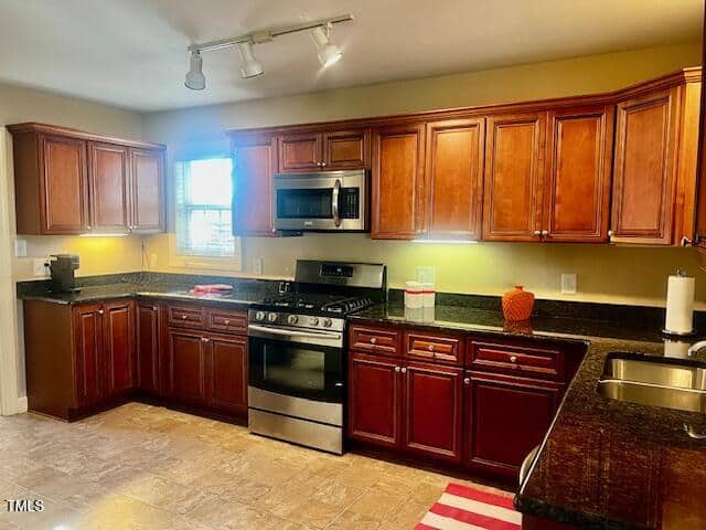 A kitchen with wooden cabinets and granite countertops. It features a stainless steel oven and microwave, a sink, and track lighting. There is a coffee maker on the counter and a red striped mat on the floor.