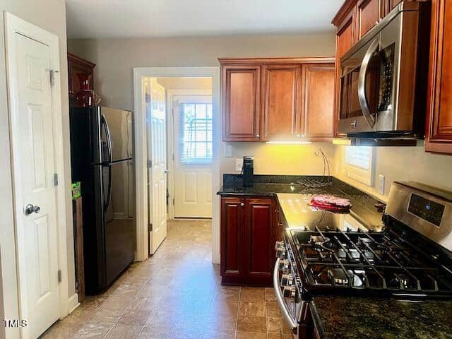 A modern kitchen with stainless steel appliances, including a refrigerator and a gas stove with an oven. Cherry wood cabinets line the walls, and a granite countertop is on the right. A window and a door are visible in the background.