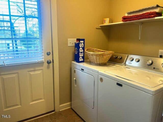 Laundry room with a washing machine and dryer under a shelf. A basket sits on the appliances, next to a Pepsi box. A window with blinds is on the left, and folded red and orange towels are on the shelf above.