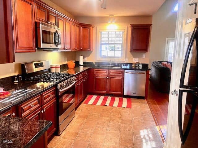 A kitchen with cherry wood cabinets, stainless steel appliances, and a black countertop. There is a gas stove, microwave, and dishwasher. The floor is tiled, and a striped red and white rug lies in front of the sink. A window is above the sink.