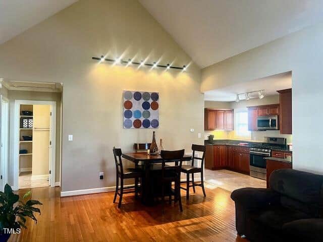 Open-concept dining and kitchen area with hardwood floors. A dining table with chairs is in front of a wall featuring a geometric artwork. The kitchen has wood cabinetry, stainless steel appliances, and recessed lighting. A plant is near the foreground.