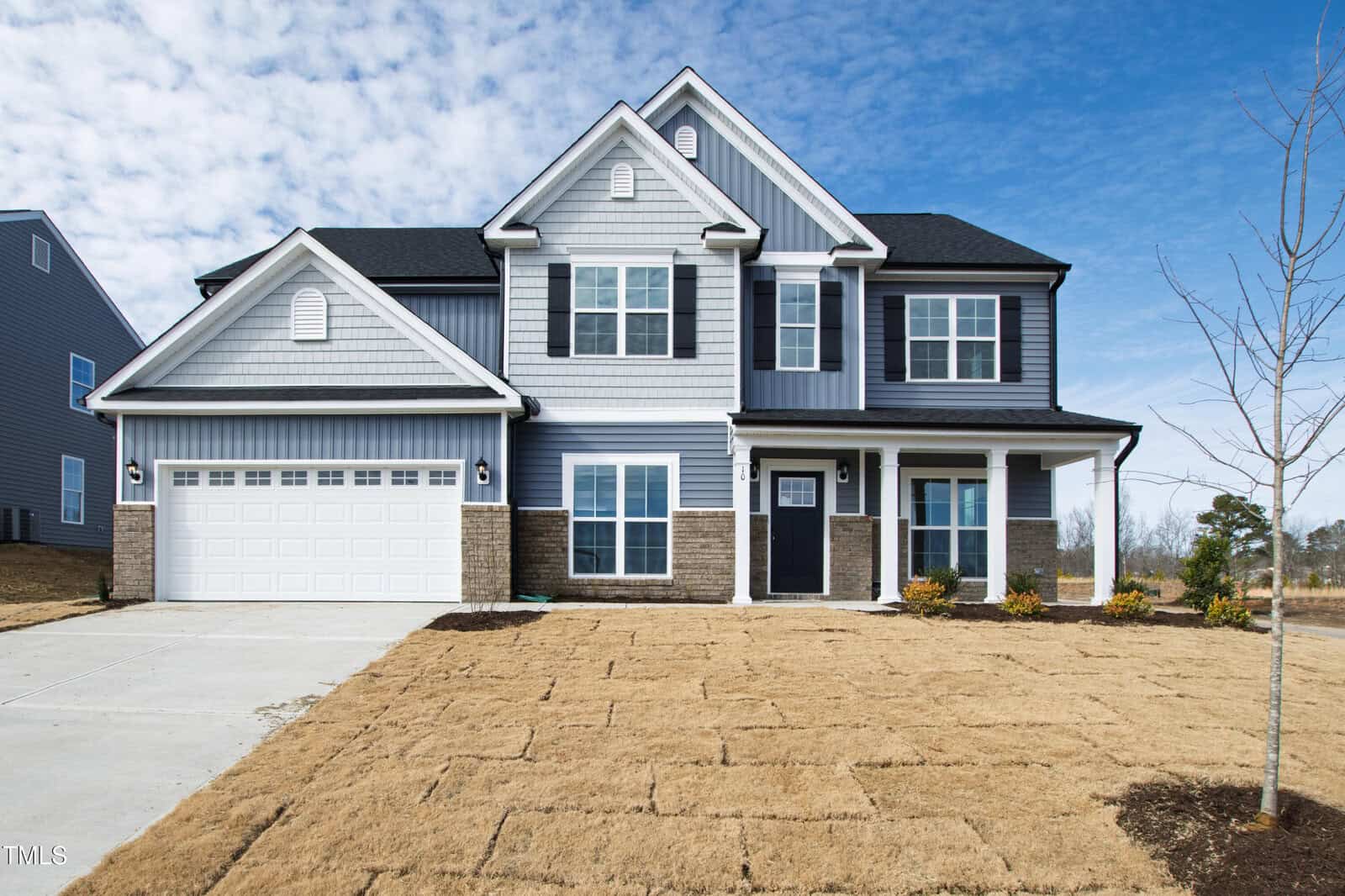 Two-story suburban house with gray siding, black shutters, and brick accents. Features a double garage, a porch, new sod in the front yard, and a clear blue sky overhead.