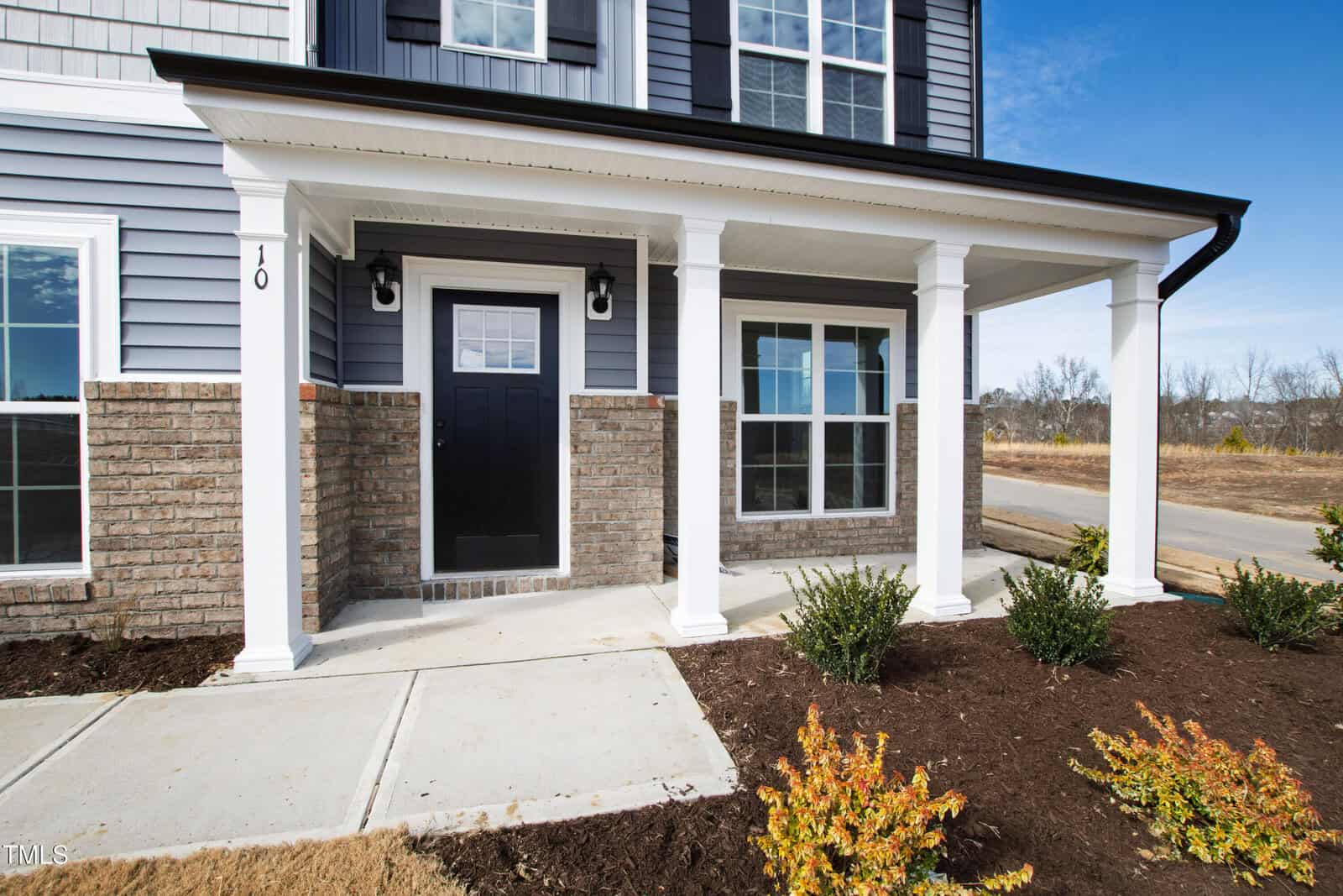 Front exterior view of a modern house with a covered entryway, black front door, white columns, brick and siding facade, two large windows, and newly planted shrubs along a concrete walkway.