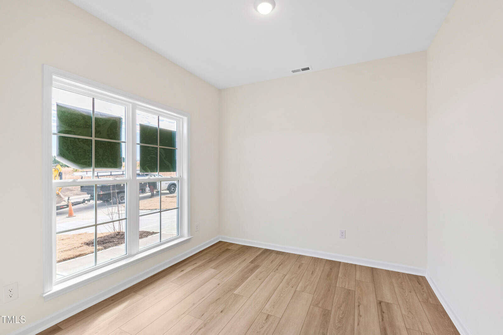 A small, empty room with light-colored walls, wood flooring, a large window letting in natural light, and a ceiling light. Some construction work is visible outside through the window.