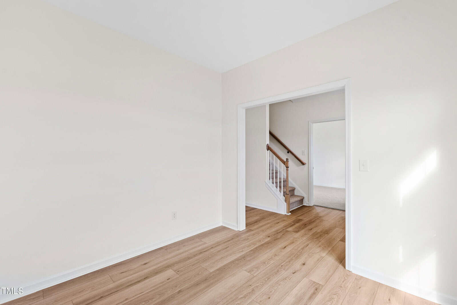 A bright, empty room with light wood flooring, off-white walls, and an open doorway leading to a carpeted stairway and another room. Sunlight is coming through an unseen window.