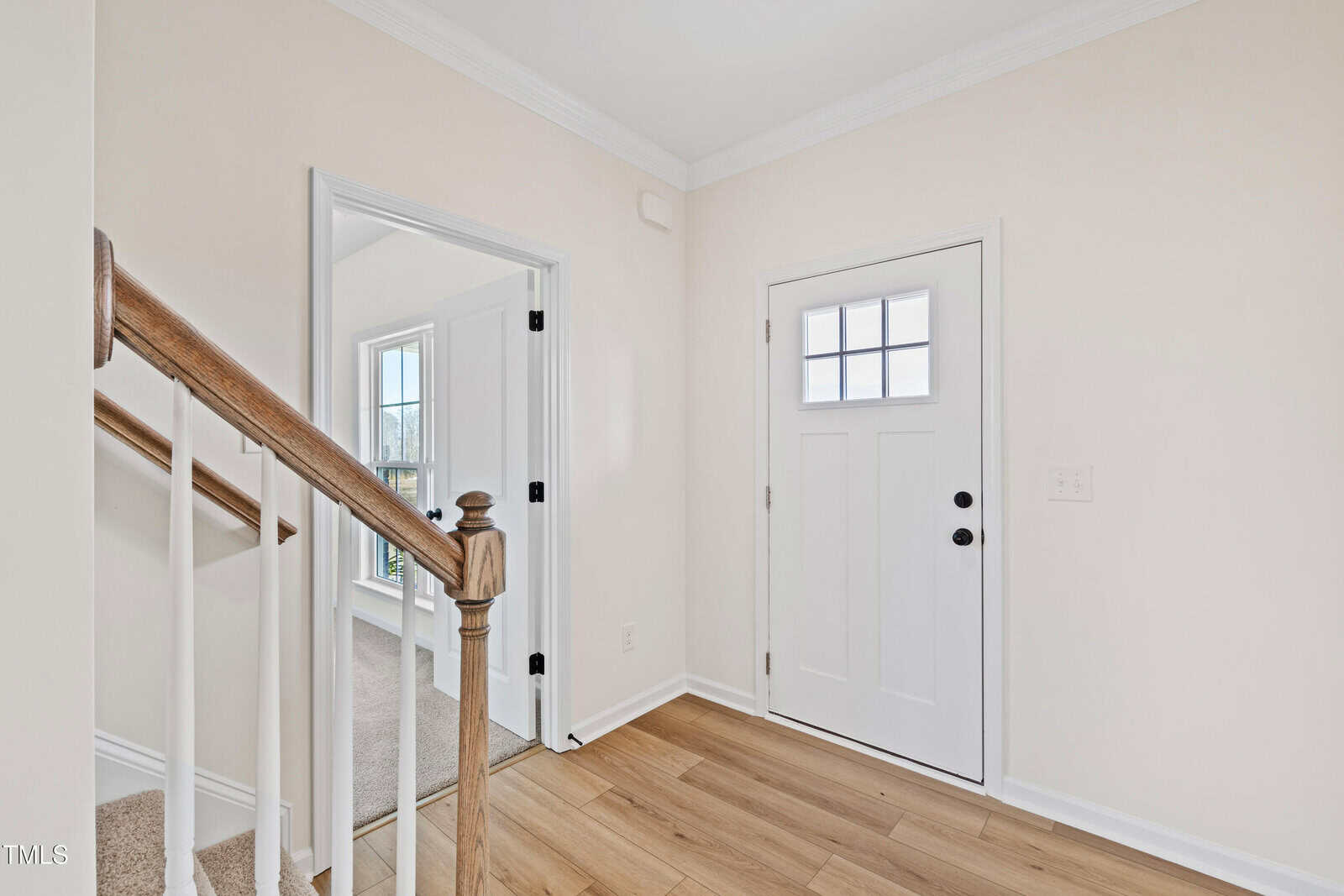 A bright entryway with light wood flooring, a white front door with glass panes, and a wooden staircase railing. An adjacent room with carpet is visible through an open doorway.