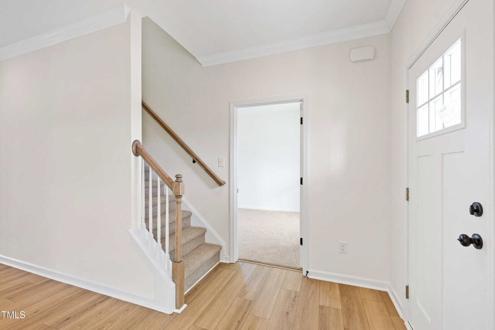 Bright entryway with light wood floors, white walls, a staircase with a wooden handrail, and a door leading to a carpeted room; white front door with glass panels on the right.