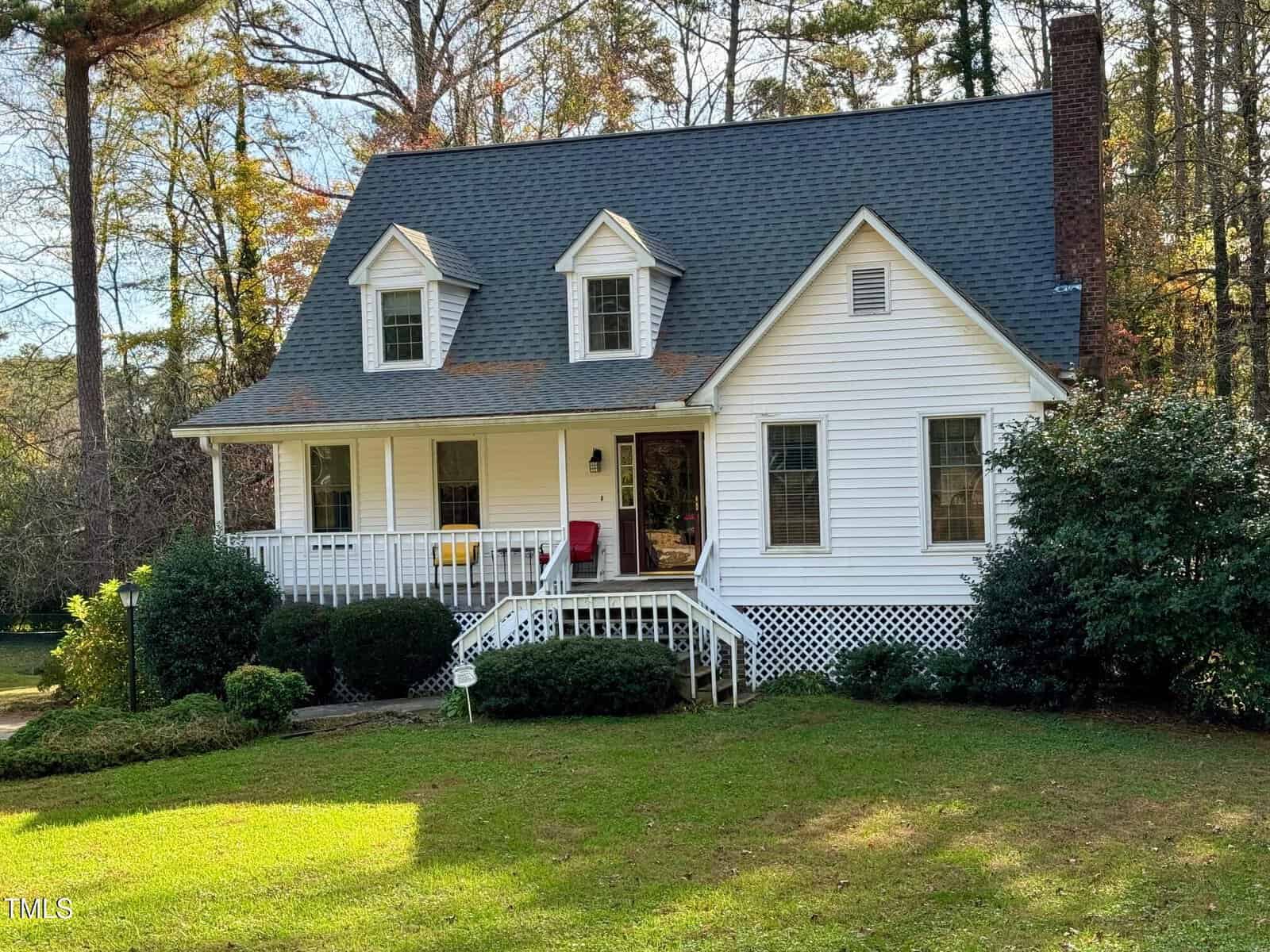 A charming white two-story house at 1613 Parker Lane, featuring a front porch, gray shingle roof, and dormer windows, is surrounded by lush green grass and bushes. Trees with autumn leaves add to the peaceful, rural setting in Henderson.