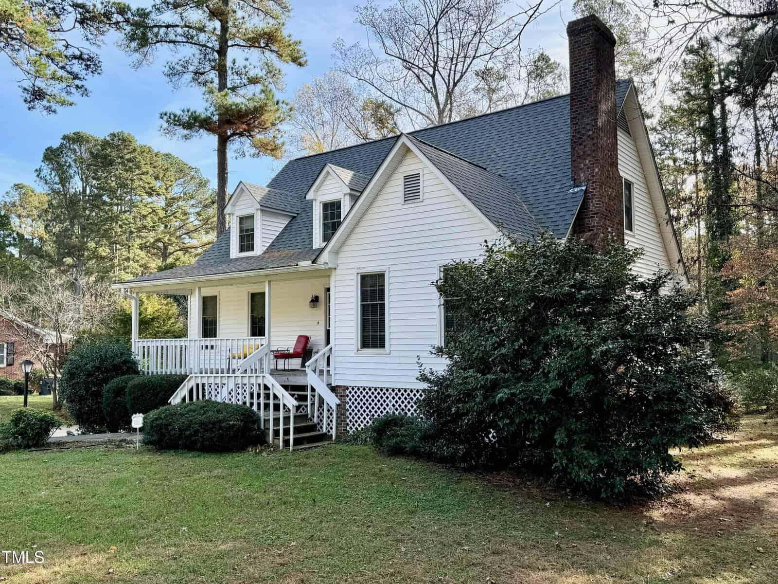 A white two-story house with a porch and a brick chimney is surrounded by trees and green shrubs. A red chair sits on the porch. The yard is grassy, and the sky is clear with a few clouds.