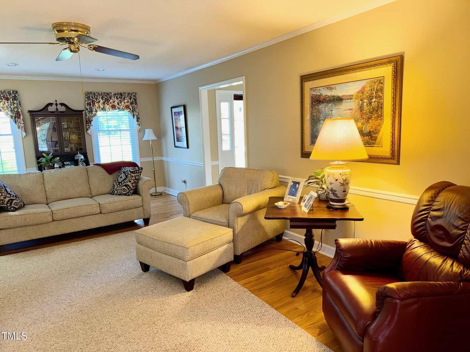 A cozy living room with beige walls and carpet, featuring a tan sofa, matching armchair with ottoman, and a brown leather recliner. A wooden side table with a lamp and framed photos sits under a landscape painting. Floral curtains adorn the windows.