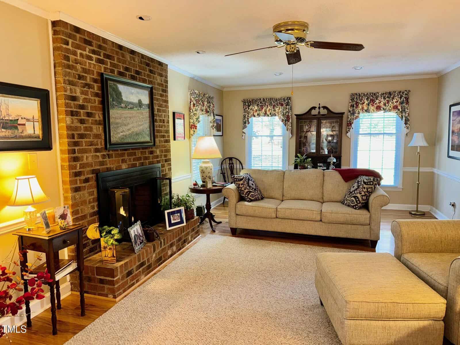Cozy living room with a beige sofa and matching ottoman, a brick fireplace, and a wooden cabinet. Framed pictures and lamps decorate the room, complementing the neutral walls and floral curtains. A ceiling fan hangs above.