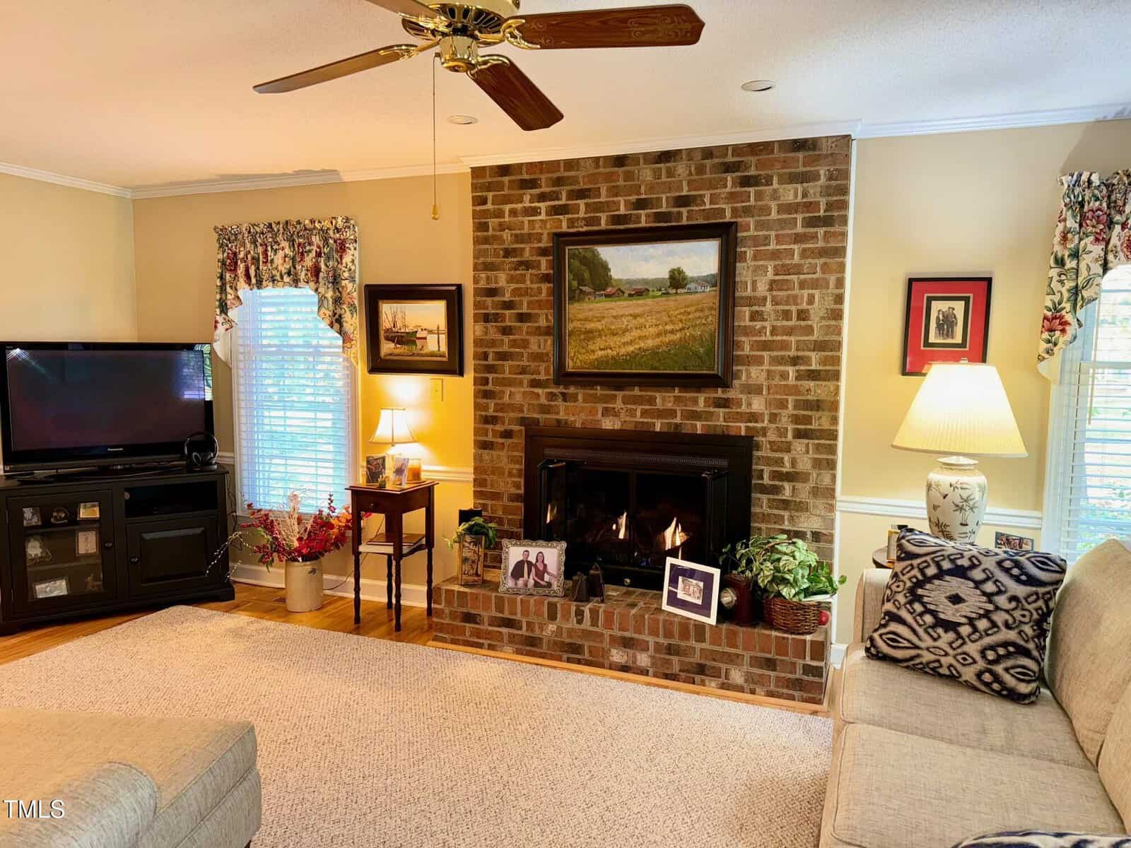 A cozy living room with a brick fireplace framed by a wooden mantel displaying family photos. A comfortable beige sofa sits nearby. A TV stands in the corner, and the room is decorated with floral curtains, lamps, and artwork on the walls.