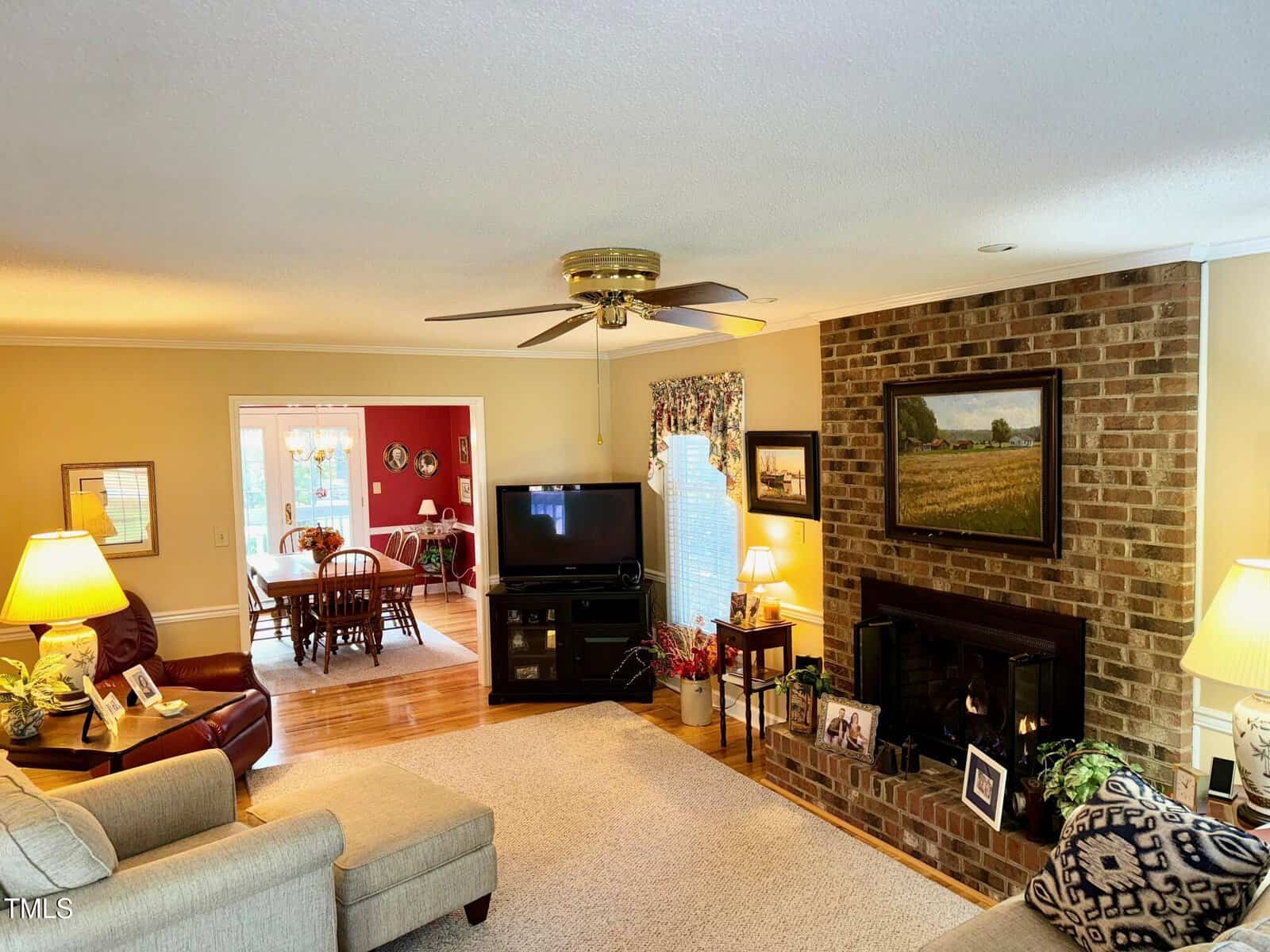 A cozy living room with beige walls and a brick fireplace featuring a painting above. The room includes a TV, sofa, armchairs, and decorative plants. A ceiling fan hangs above. An adjacent dining room can be seen through an open doorway.