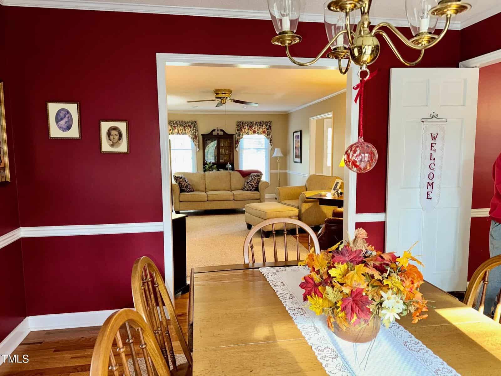 Dining room with a wooden table, autumn-themed centerpiece, and lace runner. Red walls with picture frames, and white trim. A chandelier hangs above. The room opens to a living area with beige sofas and a ceiling fan. A "Welcome" sign hangs on the door.