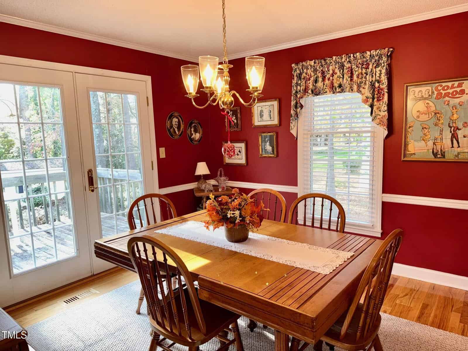 A cozy dining room with red walls is illuminated by a chandelier. A wooden table with six chairs features a floral centerpiece. A sheer curtain filters light through a window, and framed art decorates the walls. French doors lead to a sunlit deck.
