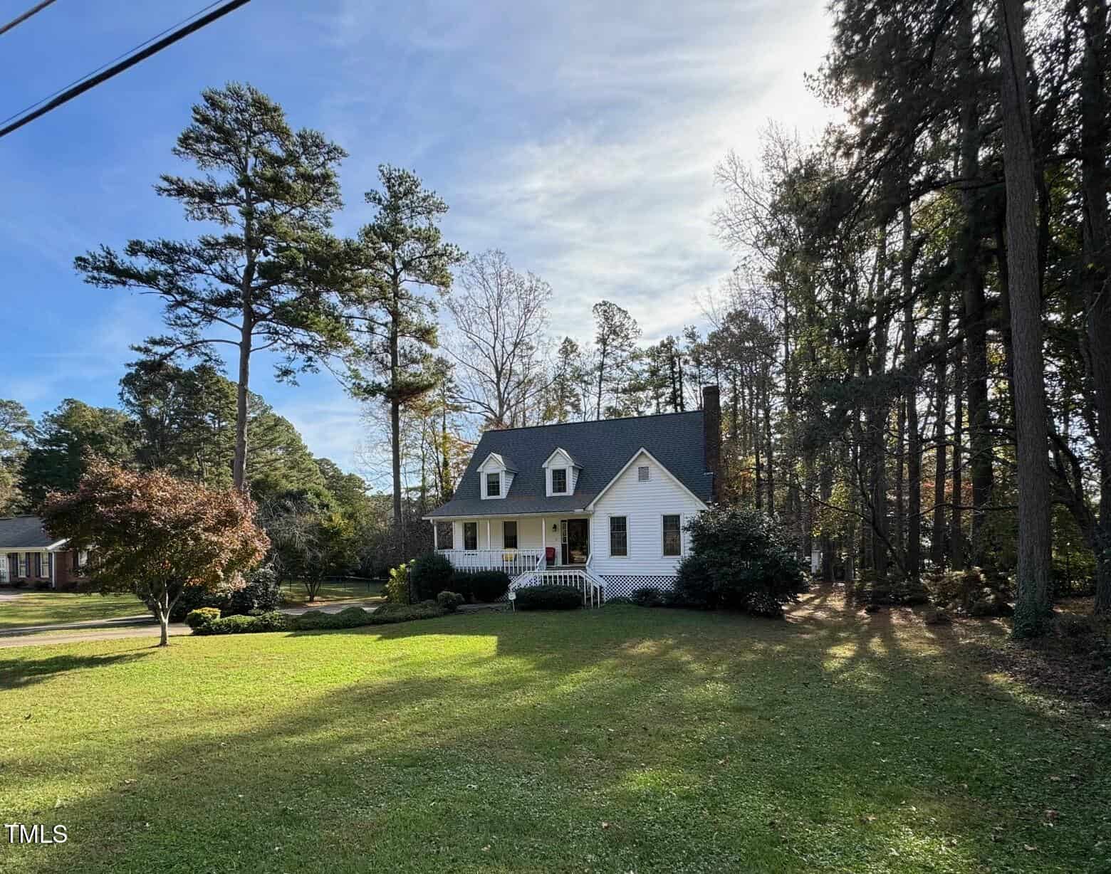 A white, two-story house with a chimney and front porch is nestled among tall trees. The house has a well-maintained lawn and a few bushes, creating a peaceful and inviting atmosphere on a sunny day.