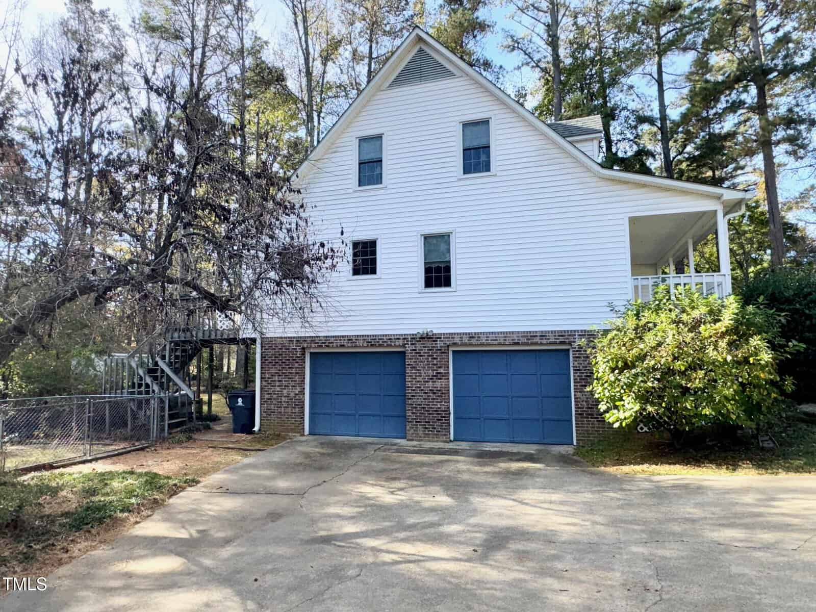 A two-story house with white siding and a brick foundation, featuring two blue garage doors. There's a porch on the second floor, trees in the background, and a chain-link fence to the side. The driveway is in the foreground.