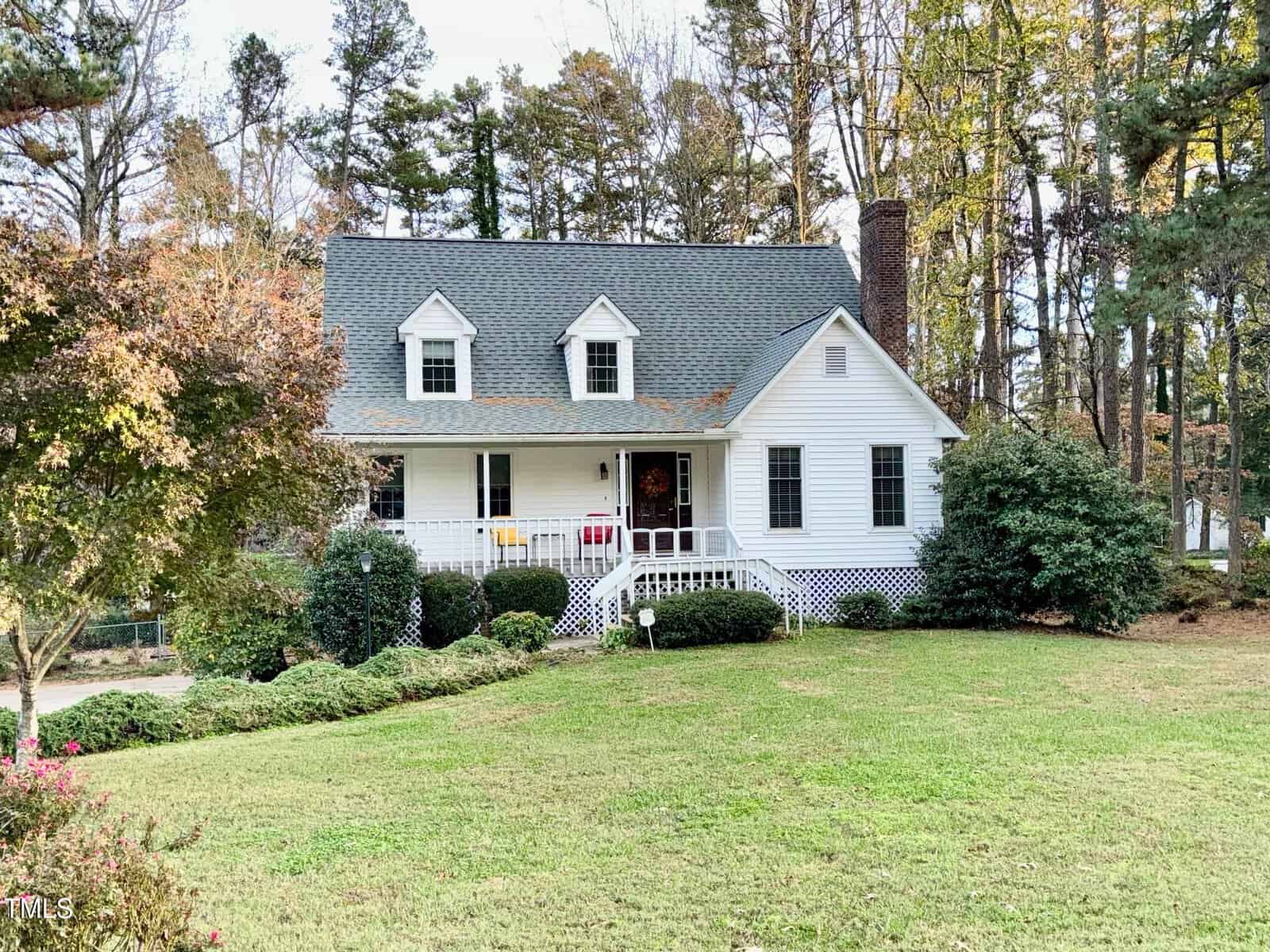 Nestled at 1613 Parker Lane, this charming white house in Henderson boasts a gabled roof and chimney, set amidst tall trees and lush greenery. The inviting front porch features a wreath, while the foreground showcases a neatly trimmed lawn.