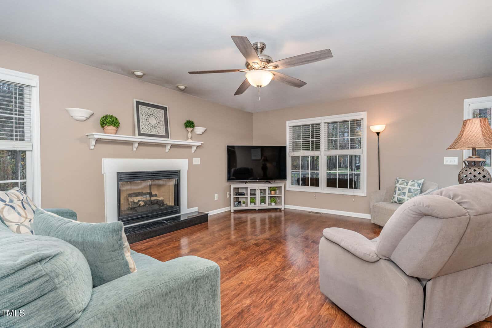A living room with hardwood floors features a gray couch, a beige recliner, and a fireplace with a white mantel. A large TV sits on a stand nearby. Decorative plants and a framed picture are on the mantel. Ceiling fan and lamp provide light.
