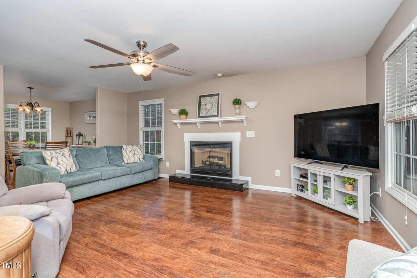 A spacious living room with hardwood floors featuring a gray couch and a recliner. A fireplace with a white mantel is centered against the far wall, and a large flat-screen TV is placed on a stand to the right. A ceiling fan hangs from the ceiling.