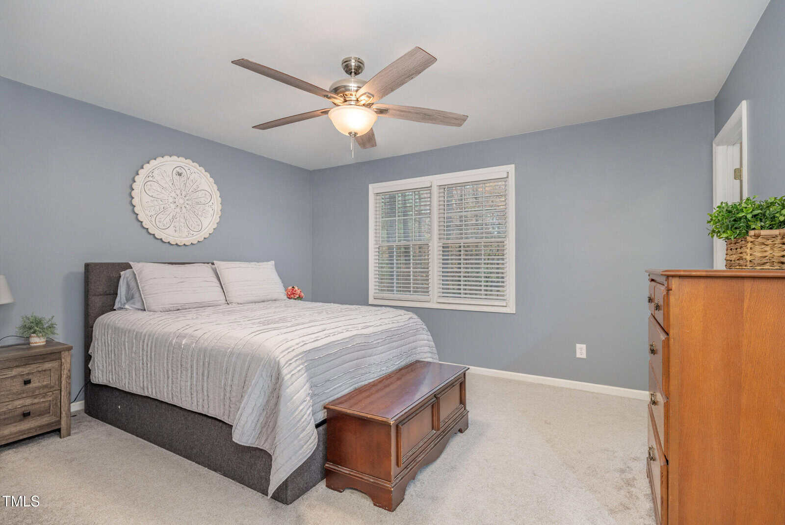 A bedroom with light gray walls, featuring a bed with gray and white bedding. A ceiling fan is overhead, and there are nightstands with lamps on each side. A wooden chest sits at the foot of the bed, and a dresser is against the wall. A window with blinds is visible.