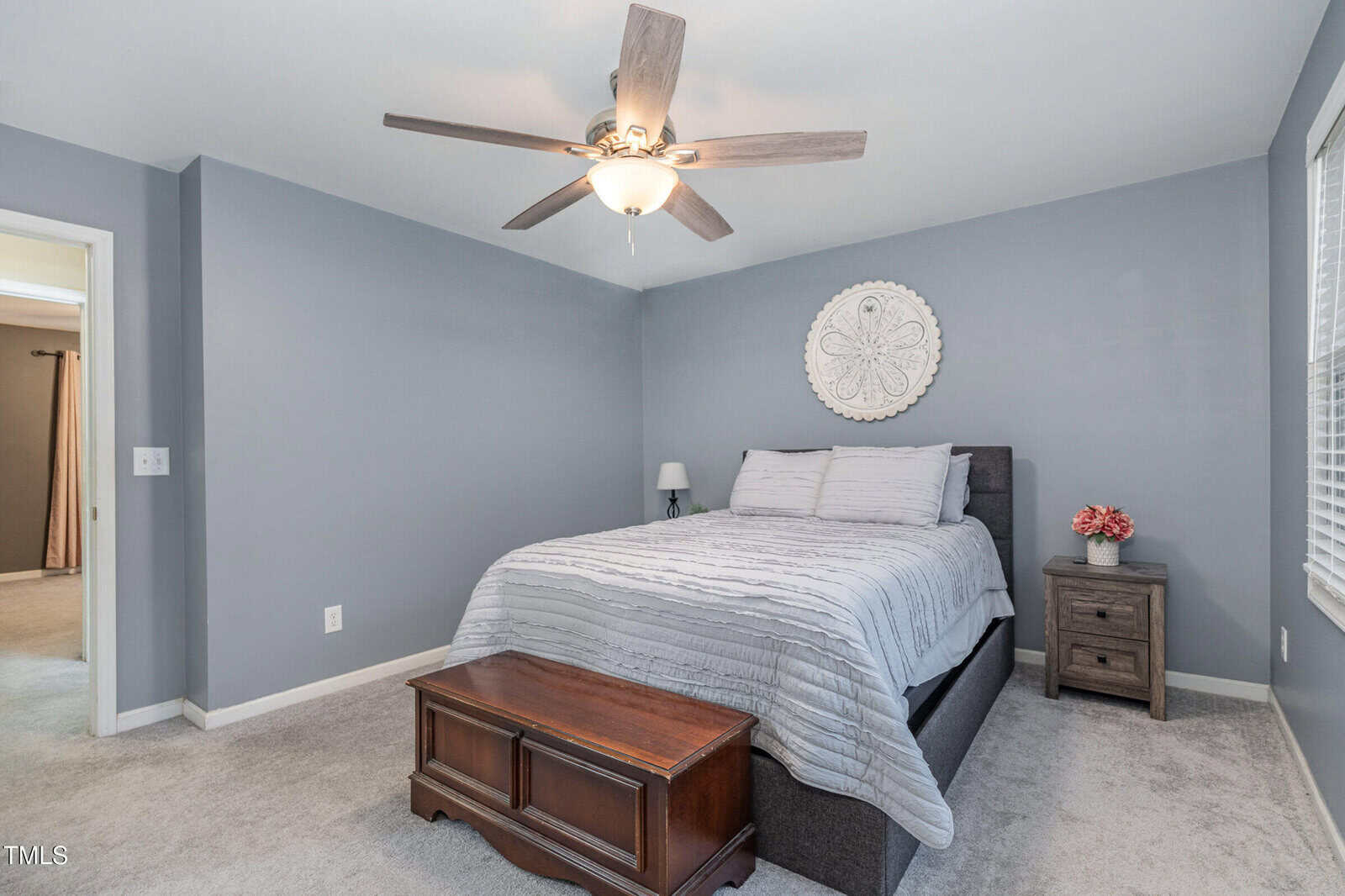A bedroom with gray walls features a double bed with a gray quilt and two pillows. Above the bed is a decorative circular wall piece. A nightstand with a lamp and flowers sits to one side. There is a ceiling fan above and a wooden chest at the bed's foot.