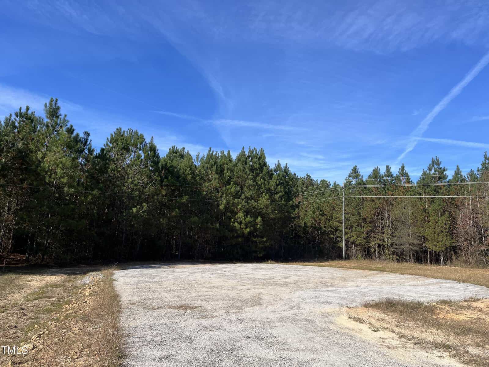 A gravel clearing is surrounded by dense pine trees under a blue sky with wispy clouds. Utility lines run across the right side of the scene. There is sparse grass along the edges of the clearing.