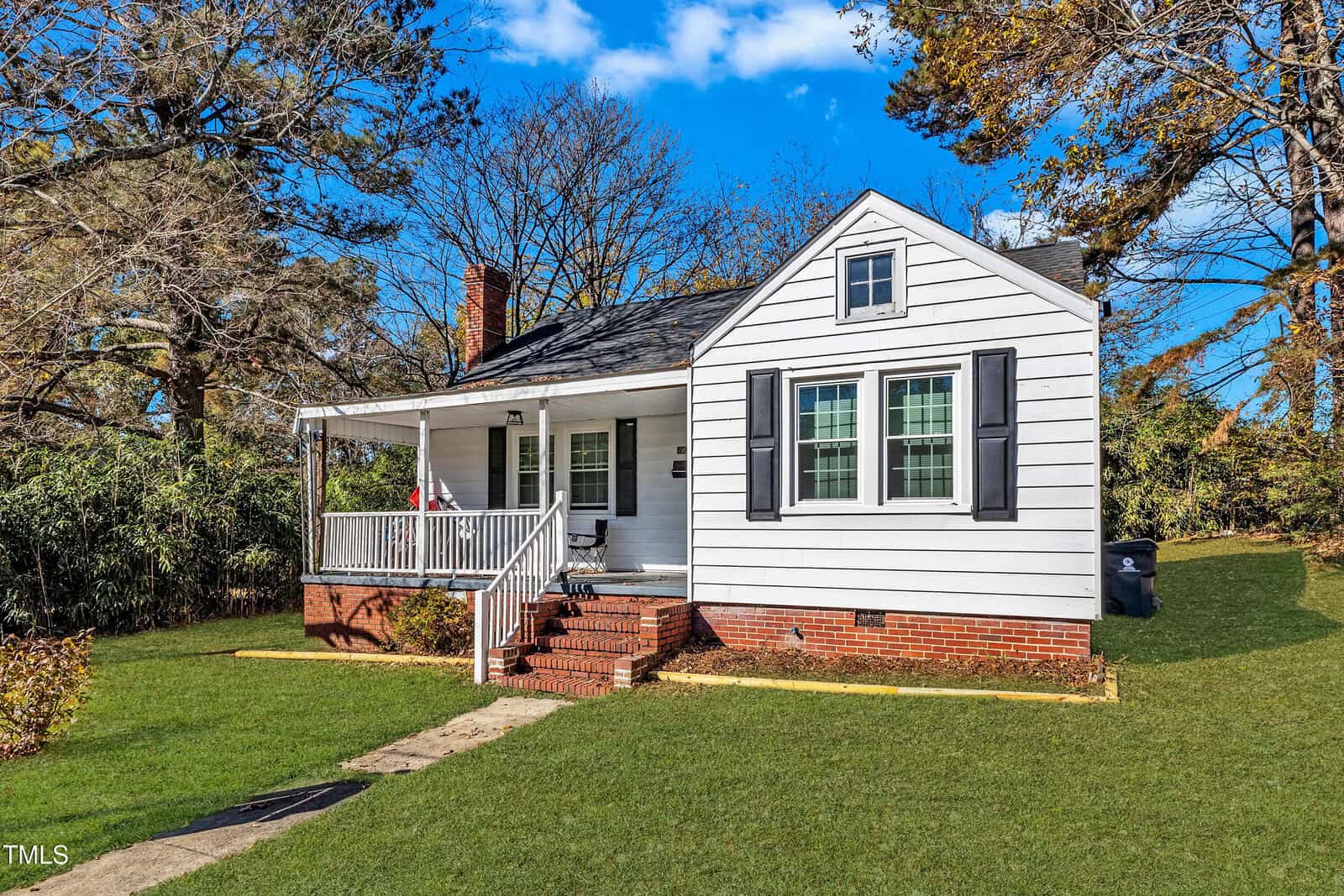The charming house at 514 Zene Street in Henderson is a white single-story home with a black roof and red brick foundation. It features a small front porch with steps and railings, nestled on a grassy lawn, with leafy trees framing the background under a clear blue sky.