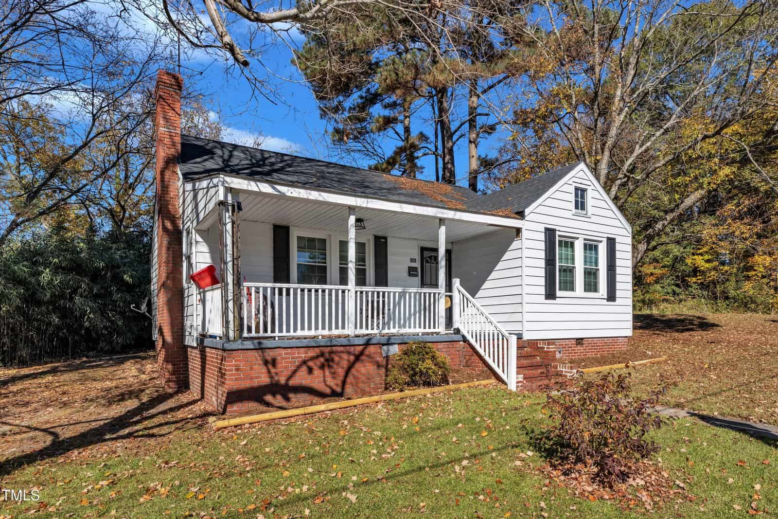 A small, white single-story house with a front porch and red brick foundation. Surrounded by autumn trees and a grassy yard, the home has a chimney on the side and a few steps leading to the porch. Blue sky and scattered clouds in the background.