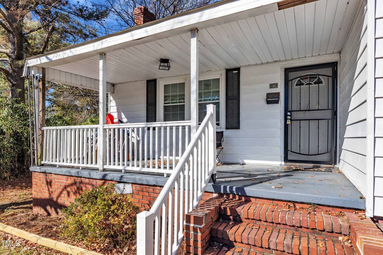 A small, white, single-story house with a covered front porch featuring a red chair. The porch has a white railing, brick steps, and a black-framed front door. Leafless trees and shrubs are visible in the background.