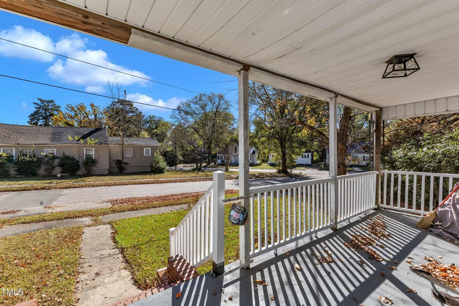A suburban street view from a covered front porch with white railings. The porch is framed by wooden beams and casts striped shadows on the floor. The neighborhood features trees with autumn leaves and single-story houses.