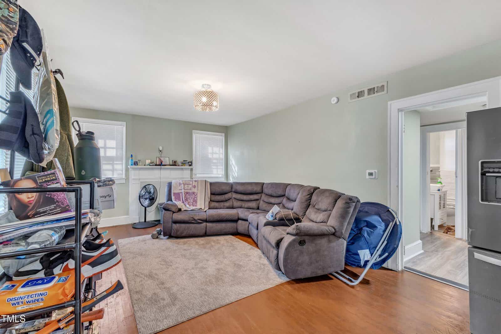 A cozy living room features a large L-shaped brown sectional sofa on a tan rug. A shelf filled with various items stands on the left, and the room is well-lit with a modern ceiling light. A doorway leads to an adjoining room on the right.