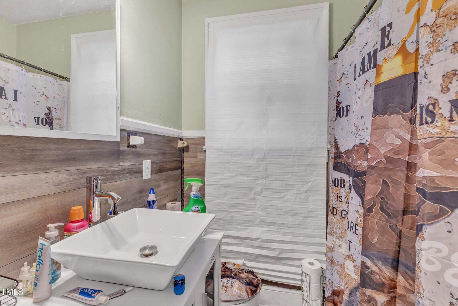 A bathroom with a modern white sink and faucet on a countertop. Various cleaning supplies are placed around the sink. The shower curtain has a decorative motif, and a partially open window with a white blind is in the background.