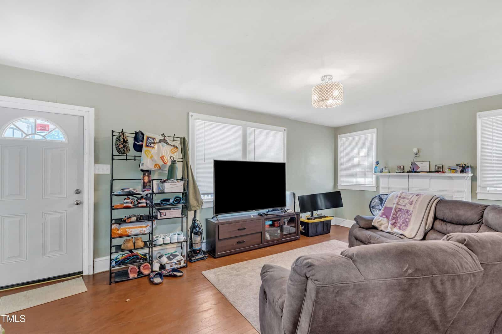 A cozy living room features a gray recliner, wall-mounted TV, and a shelf with shoes and hats. A white door with a small window is on the left, and a decorative light fixture hangs from the ceiling. Blinds cover the windows, letting in natural light.