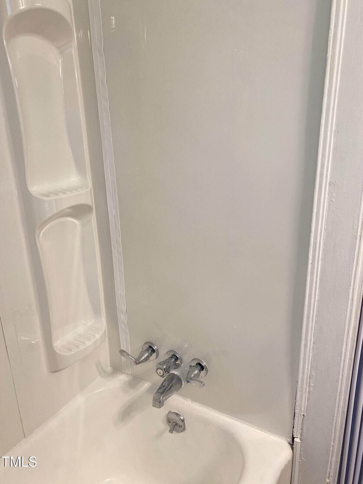 Close-up of a white bathtub with a built-in shower and three wall-mounted knobs. The bathtub has an integrated shelf for toiletries.