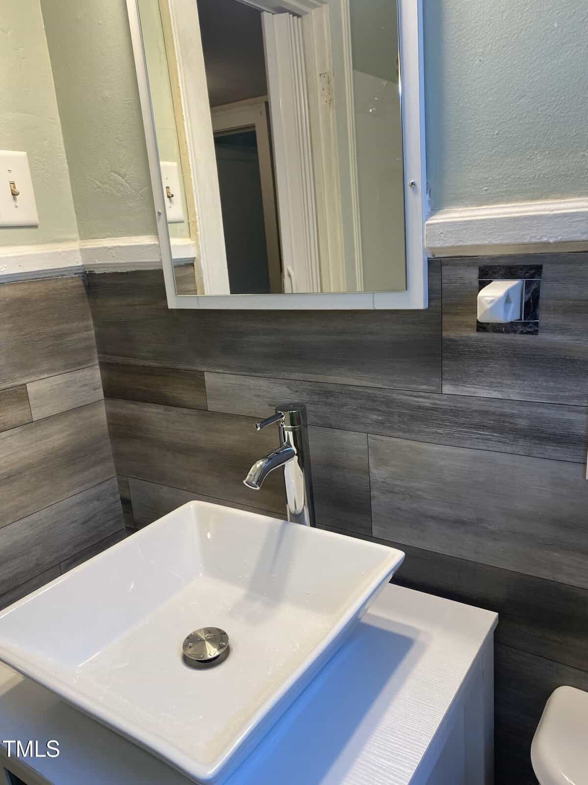 A modern bathroom with a square white vessel sink and chrome faucet set against a backsplash of gray wooden tiles. Above the sink is a rectangular mirror with a white frame. The walls are painted light green.