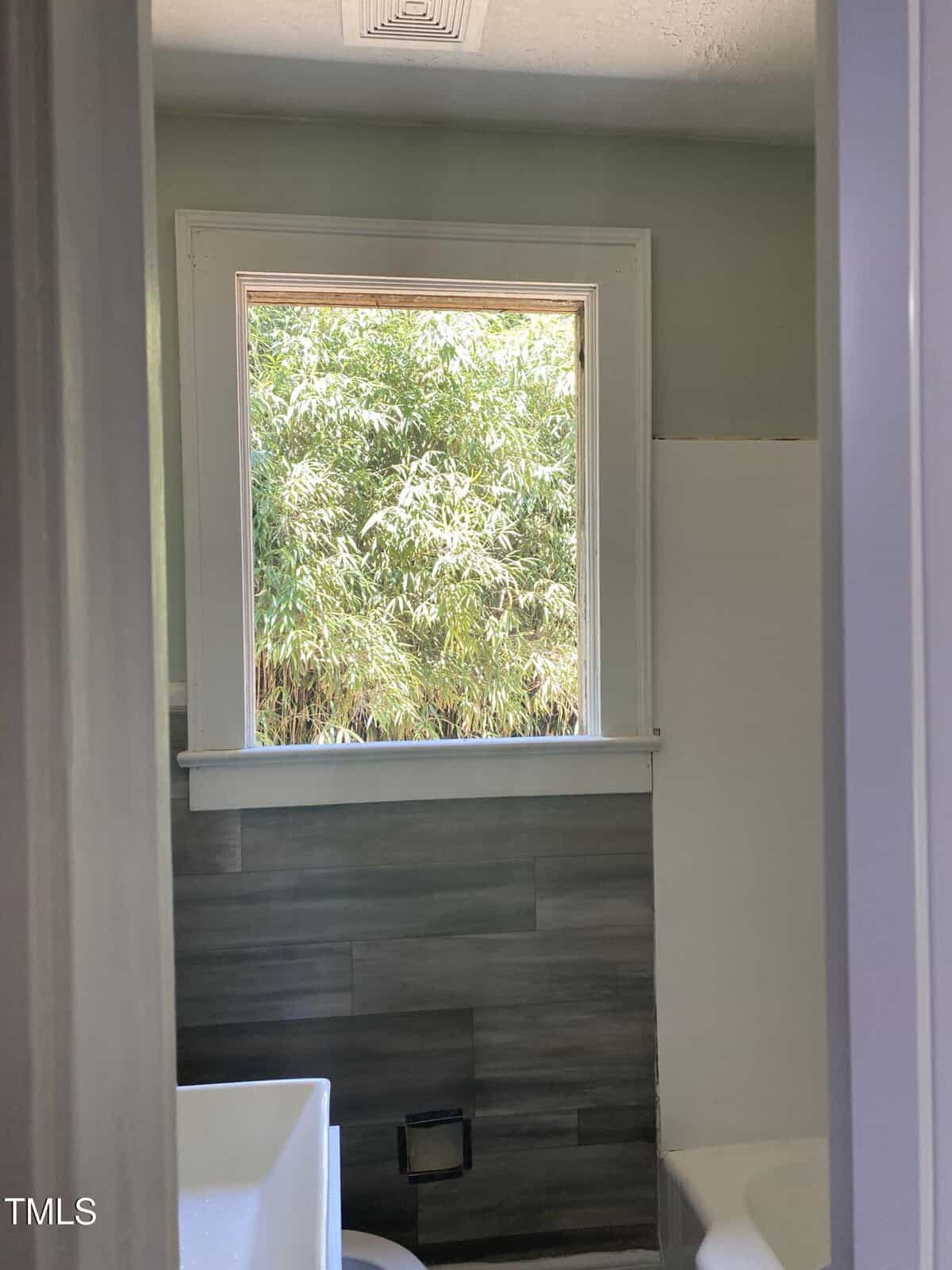 A small bathroom with a window showing dense green foliage outside. The room features gray wall tiles, a white bathtub partially visible on the right, and a white sink on the left. Sunlight streams in through the window, illuminating the space.