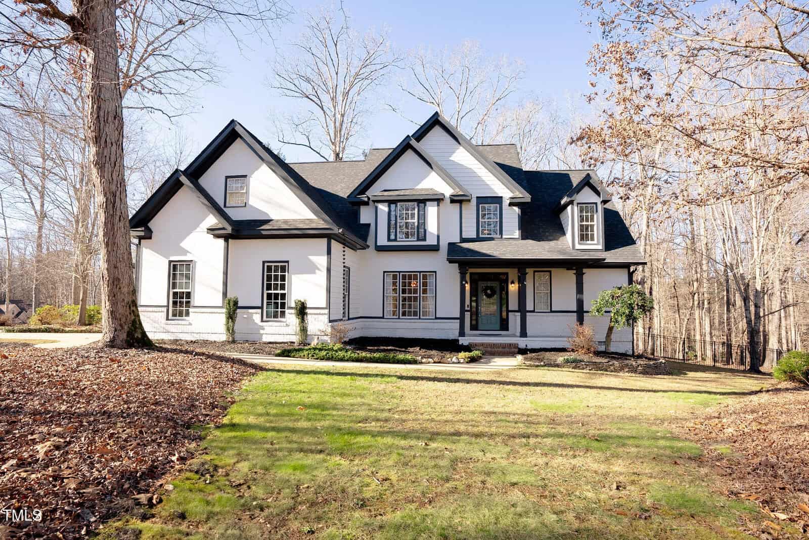 A two-story white house with black accents on 4022 Fernbank Way, Youngsville, features a steep gabled roof and covered front porch, nestled among bare trees and a partially grassy yard in a peaceful wooded area.